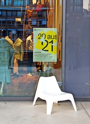 Ein weißer Stuhl steht vor einer Glaswand an der das 20 aus 21 Plakat hängt