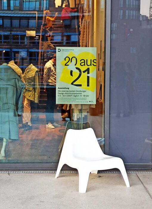 Ein weißer Stuhl steht vor einer Glaswand an der das 20 aus 21 Plakat hängt