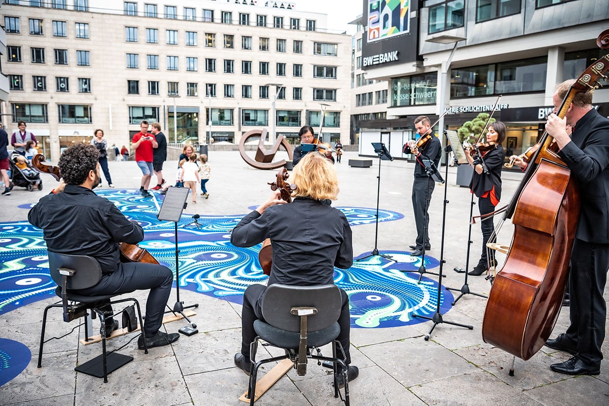 Ein Orchester sitzt mit dem rücken zur Kamera vor einem Kunstwerk am Boden