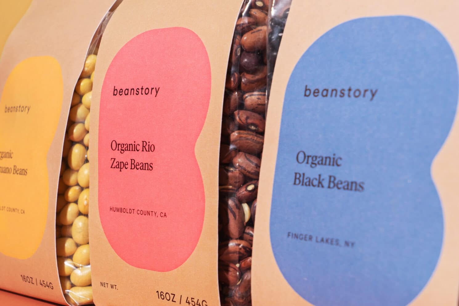 Visual Identity für den Bohnenvertrieb Beanstory: Details des Packaging Design drei verschiedener Bohnensorten. Auf der Papierverpackung ist im Hintergrund immer das B in Bohnenform zu sehen, aber für jede Sorte eine andere Farbe: Links in Gelb für Peruano Bohnen, in der Mitte rosa für Zape Bohnen und rechts blau für Schwarze Bohnen.