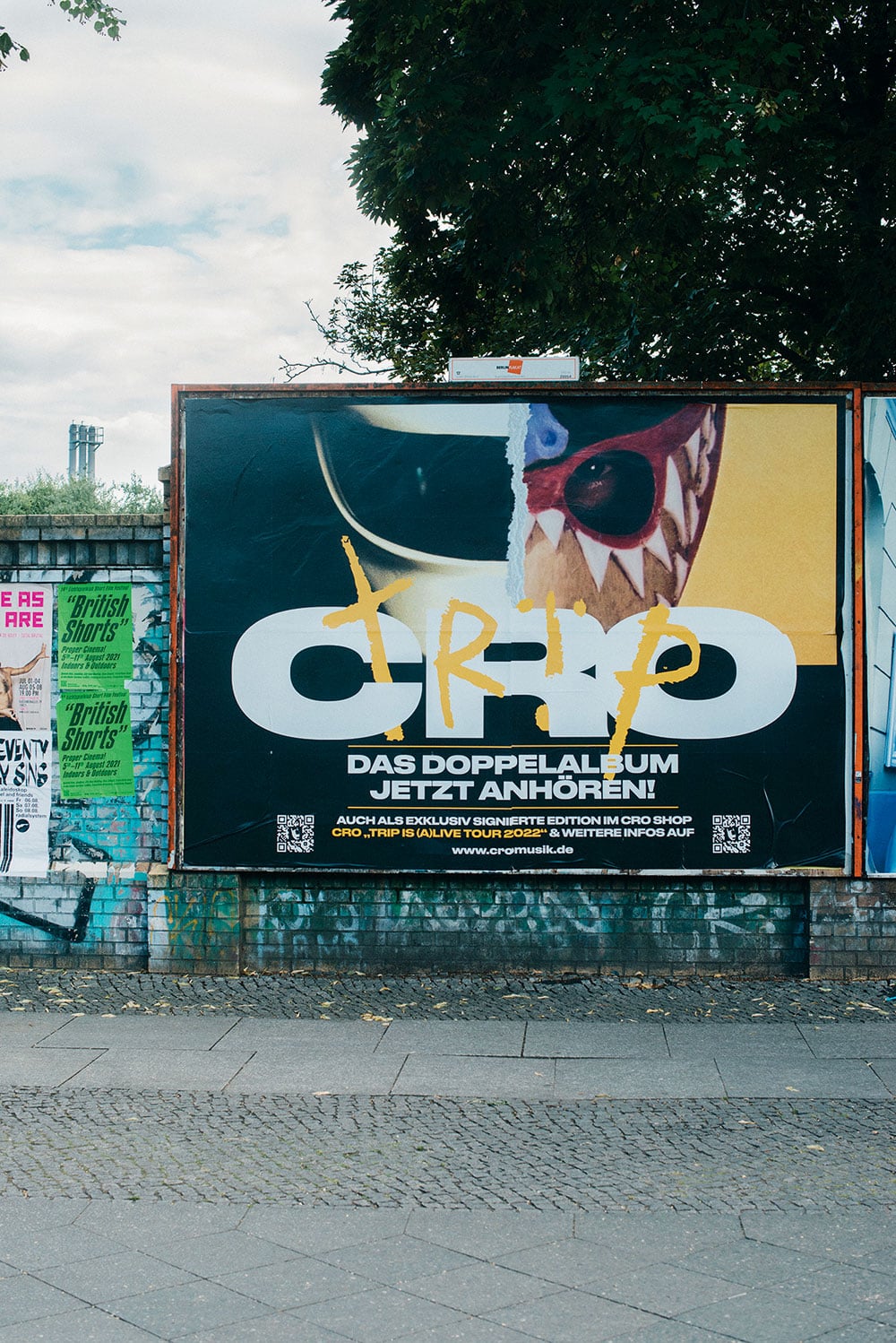 Plakat für das Co-Album »Trip«, das auf eine Plakatwand plakatiert wurde. Das Poster zeigt den Kopf des Rappers Cross von zwei unterschiedlichen Masken bedeckt. 