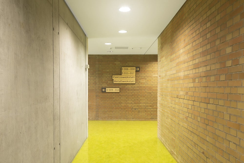 Flur im Gebäude »D« der FOSBOS. Die Wände sind zum Teil gelb-orange gemauert, zum Teil aus Beton. Der Boden ist knallgelb. Das Leitsystem weist den Weg zu unterschiedlichen Räumen und Werkstätten. 