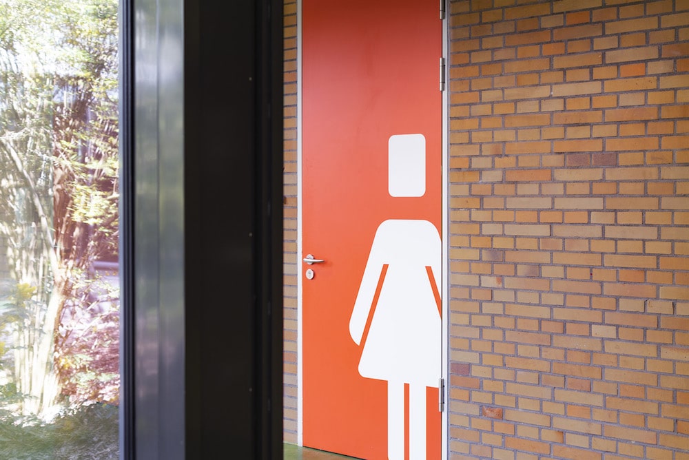 Toilettentür gestrichen in kräftigem orange und mit dem weißen Icon einer Person in Kleid.
