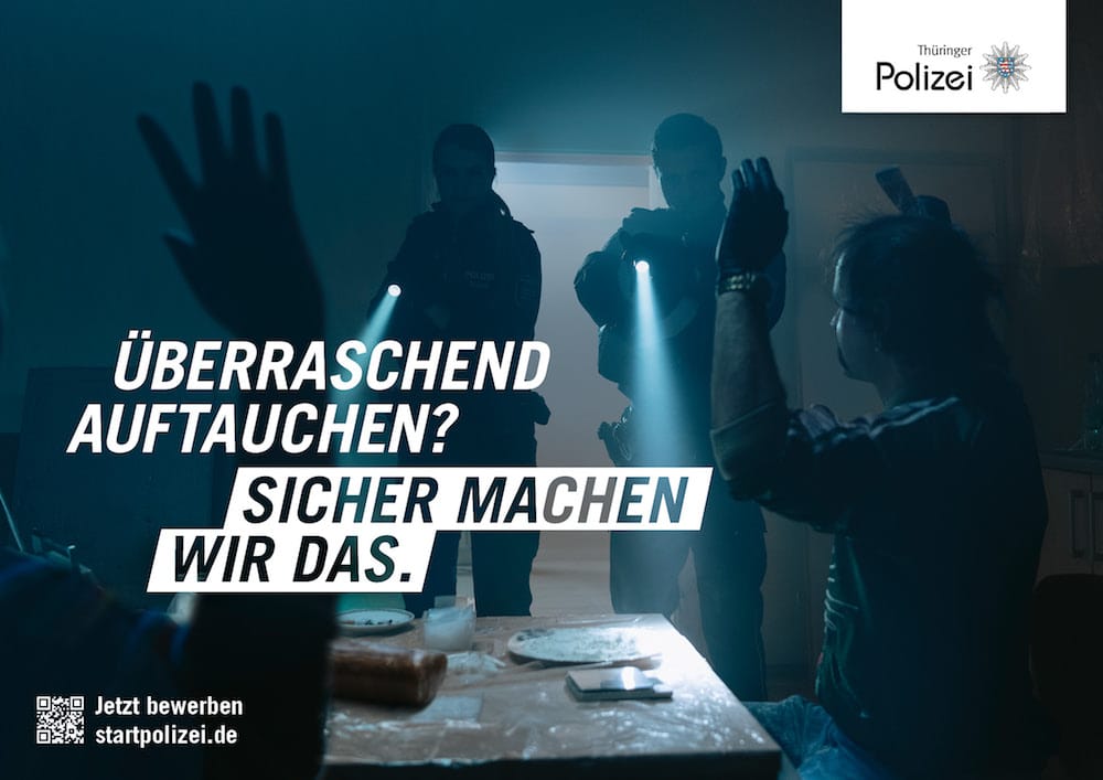 Recruitingkampagne für die Thüringer Polizei: Beamt:innen mit Taschenlampen zielen mit ihren Waffen auf mehrere Personen an einem Tisch, die die Hände heben. Auf dem Tisch sind vermutlich Drogen zu sehen. Zu lesen: »Überraschend auftauchen? Sicher machen wir das.«