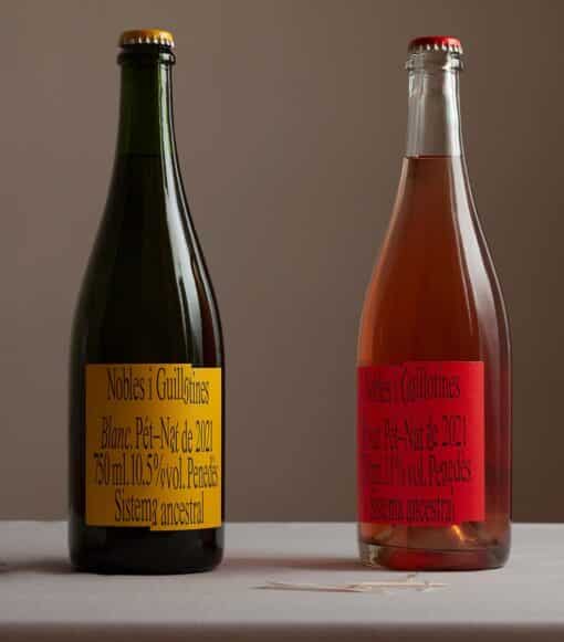 Zwei Weinflaschen stehen nebeneinander vor neutralem Hintergrund. Die dunklere, links trägt ein hellgelbes Label das in zwei Teile geschnitten ist. Die hellere Flasche mit Rosé, recht, trägt ein rotes label