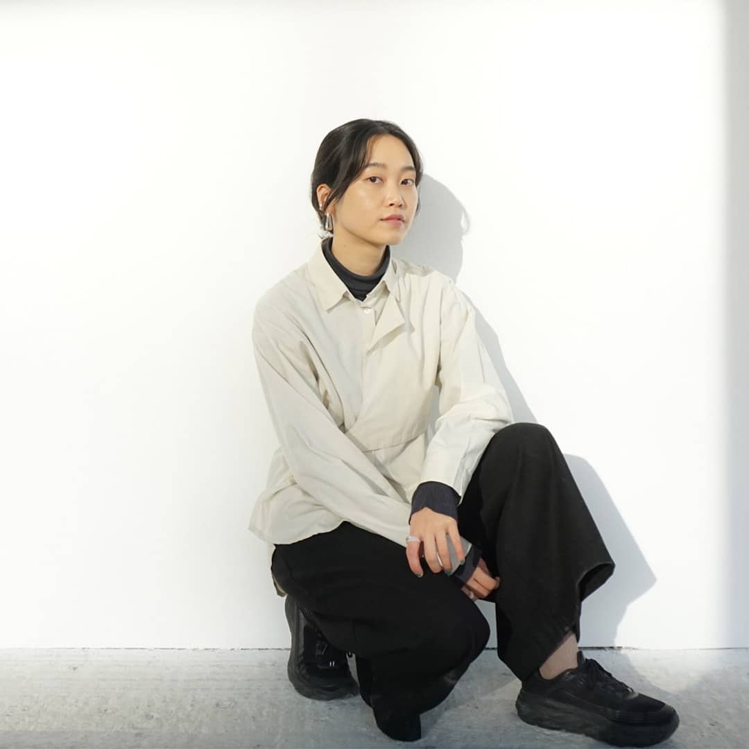 Portät von Jeongkyoung Woo: Die Künstlerin hockt vor einer weißen Wand und schaut in die Kamera