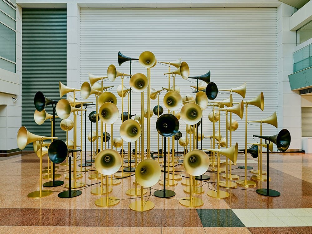 Depicted is the artwork »Sound Cloud« by Yuri Suzuki at Haneda Airport in Tokio. The artwork consists of numerous trumpet like speakers that are mostly in gold and sometimes black. The speakers are each mounted on thin bars in the same color as the trumped color. 