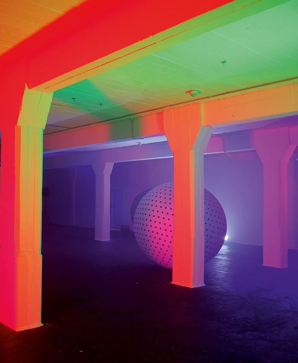 Artwork by Yuri Suzuki called »Synthesizer Playground«. Depicted is a room with white walls and columns and a dark ground, maybe a warehouse or a parking house. There are two inflatable spheres with polkadots in this room between the columns. Differently colored light is coming from unknown light sources
