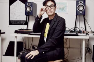 Portrait of Yuri Suzuki in his Studio, sitting on a stool, looking straight into the camera with one hand on his knee and the other one next to his face, fixing his glasses