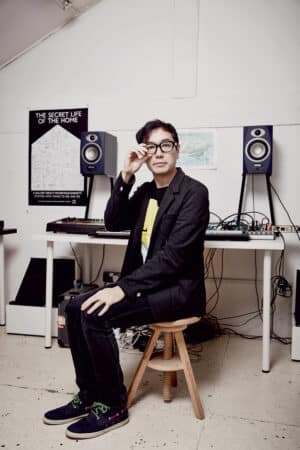 Portrait of Yuri Suzuki in his Studio, sitting on a stool, looking straight into the camera with one hand on his knee and the other one next to his face, fixing his glasses