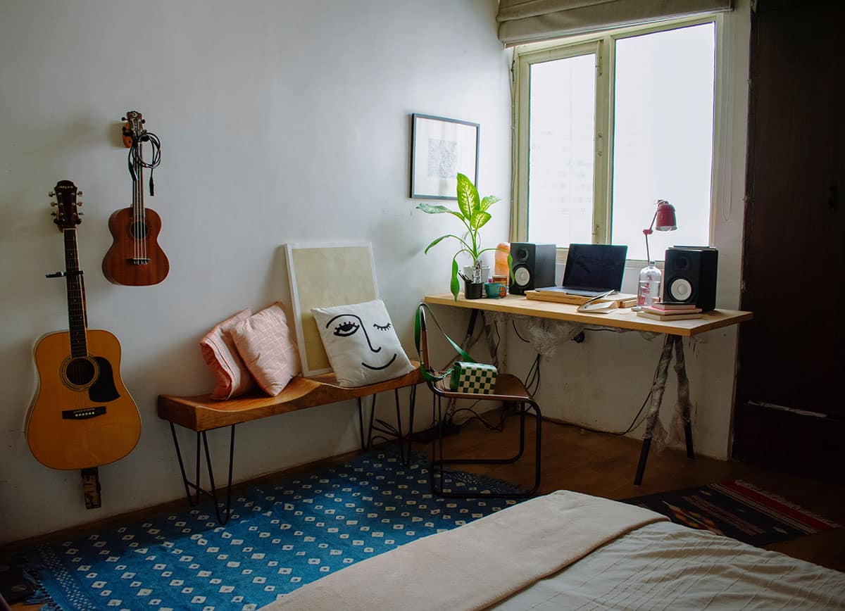 Ein Foto von Khyati Trehans Arbeitszimmer mit einem Schreibtisch am Fenster, zwei Gitarren, die an der Wand hängen und einigen Pflanzen, sowie einer Sitzbank mit Kissen