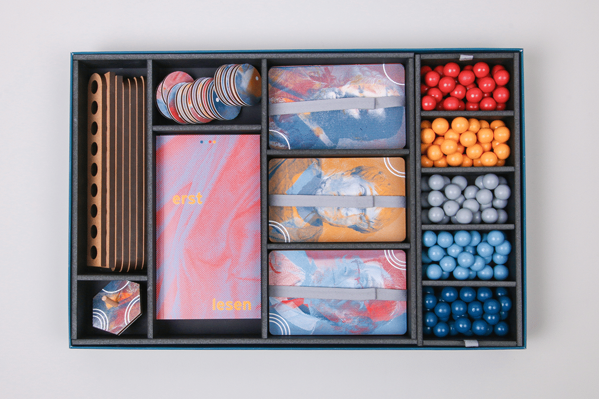 Eine Draufsicht auf den Spielkarton, in dem farbige Kugeln, drei Kartenstacks, sowie vier Ethikhefte und einige Spielmünzen liegen