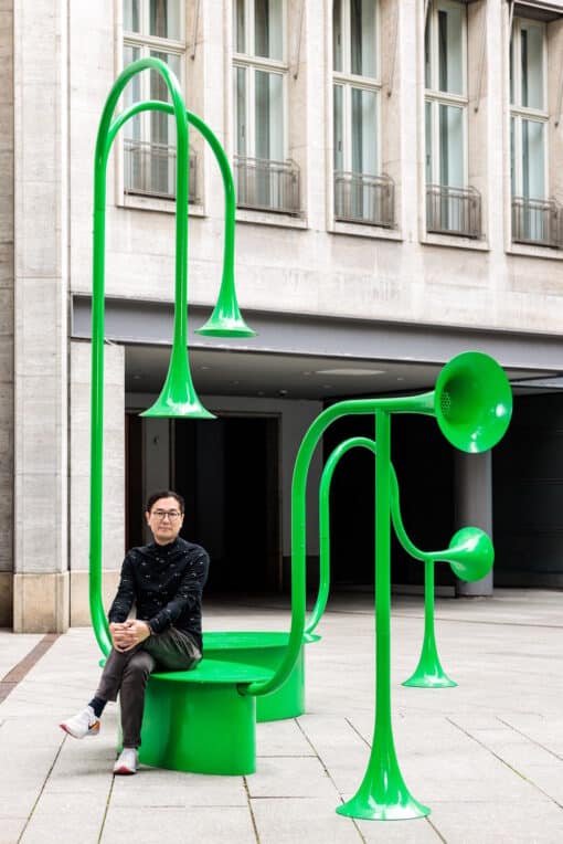 Yuri Suziki sitzt mit knallgrüner Soundskulptur mit großen Tröten vor Wohnhaus