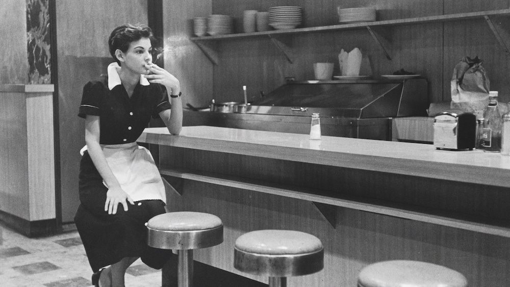 Historisches Foto einer Kellnerin, die an einem Tresen sitzt und raucht