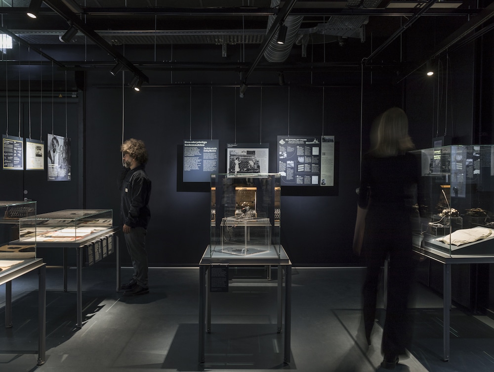 Ausstellungsdesign für das »Enigma – Museum for post, tele og kommunikation« von Atelier Brückner: dunkle Raumgestaltung mit erleuchteten Tischvitrinen und Infotafel