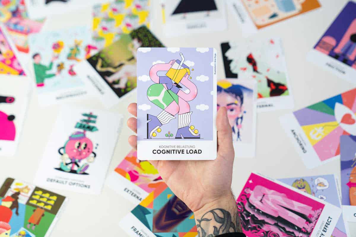 Eine Hand hält eine Illustrierte Spielkarte vor einem Tisch mit dem restlichen Kartendeck. Darauf ist eine Illustration in hellen Pastelltönen zu sehen