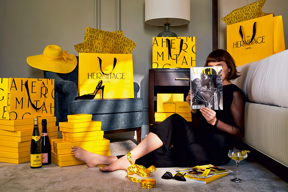 Hotelbranding: Eine Person sitzt auf einem Teppich, lehnt an einem Bett und blättert in einer Vogue. Um sie herum stehen viele gelbe Tüten mit dem Hermitage-Branding