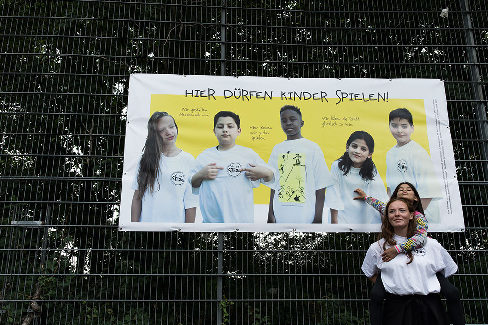 Eine junge Frau trägt ein Kind auf dem Rücken. Sie stehen vor einem Plakat, auf dem mehrere Kinder abgebildet sind. Darüber steht "Hier dürfen Kinder spielen!"
