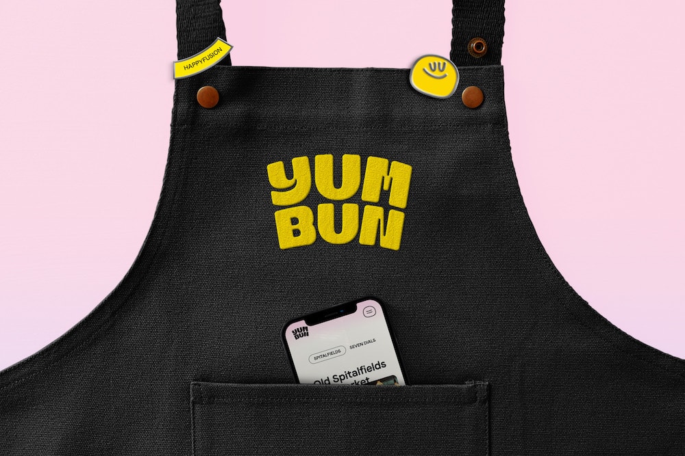 Branding für Yum Bun: Schwarze Schürze mit gewebtem Wortlogo und Smiley-Pin