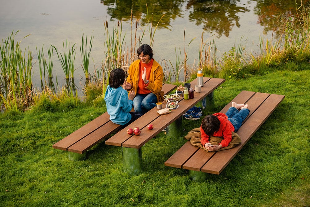 Eine Familie sitzt and einem See auf Bänken und an einem Tisch aus Holz. Die Bänke und der Tisch sind auf Beinen aus einem Material, dass die umliegende Wiese spiegelt. Daher sieht es so aus, als würden Bank und Tisch schweben.