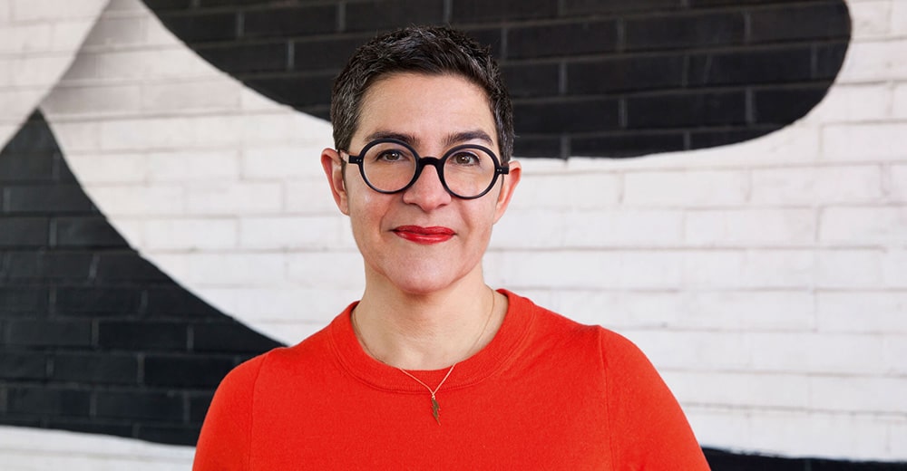 Porträtbild von Sarah Durham in einem roten Pullover vor schwarz-weißer Wand