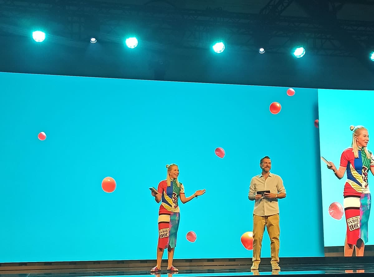 Die beiden Moderator:innen stehen vor einem blau erleuchteten Hintergrund auf der Bühne