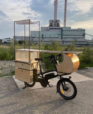 Ein Fahrrad mit einem Aufsatz, der aussieht wie eine Bar steht vor einer Fabrik auf einem Grünstreifen