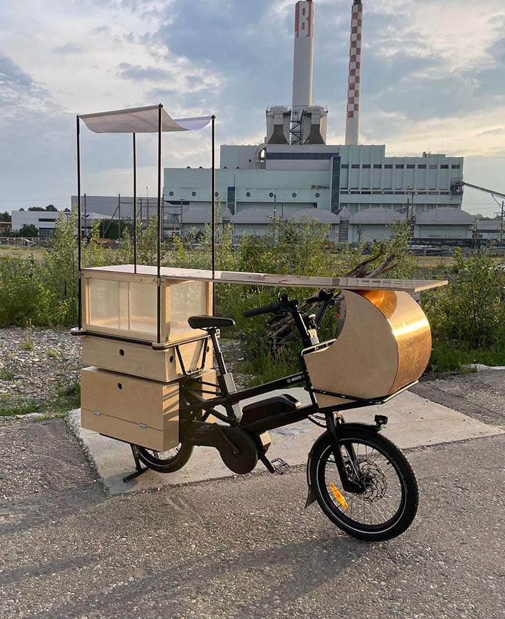 Ein Fahrrad mit einem Aufsatz, der aussieht wie eine Bar steht vor einer Fabrik auf einem Grünstreifen