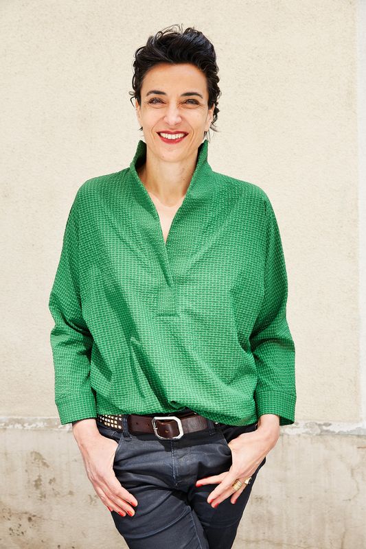 Eine Frau mit Kurzen Haaren, die in eine grüne Bluse gekleidet ist und in die Kamera Lächelt