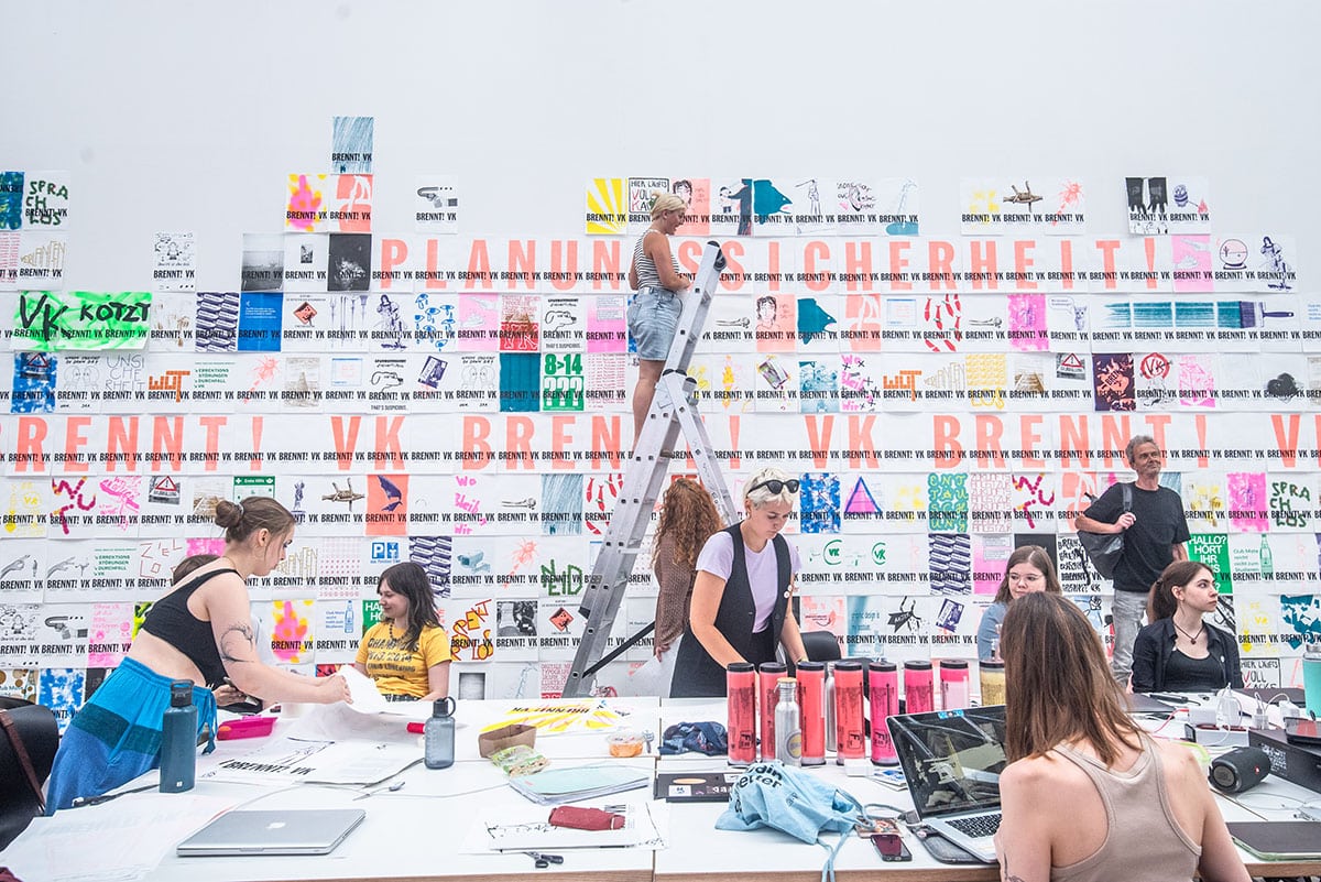 Studierende kleben mit einer Leiter Plakate an eine helle Wand. Davor arbeitet eine Gruppe an neuen Entwürfen auf einem Tisch