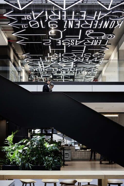 Eine Installation aus Leuchtzeichen ist an einer dunklen Decke angebracht Darunter geht ein Mann eine schwarze Treppe hinunter