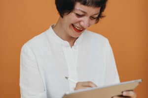 Eine fröhliche Frau mit Bob-Haarschnitt konzentriert sich darauf, mit einem Stylus auf einem Tablet zu schreiben. Sie trägt ein knackiges weißes Hemd und dunkle Hosen, mit einem warmen, sanften Lächeln auf dem Gesicht, das Amüsement oder angenehme Gedanken anzeigt. Der Hintergrund ist einfarbig orange und bietet eine warme und lebendige Atmosphäre, die zu ihrem freudigen Ausdruck passt.