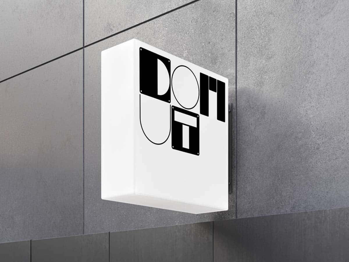Ein quadratisches, beleuchtetes Schild mit dem Schriftzug 'DONUT SHOP' in schwarz-weißem Design, das an einer grauen Wand befestigt ist