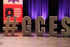 Das Bild zeigt große, dreidimensionale Buchstaben, die zusammen "#GCES" ergeben, auf einer Bühne mit einem violetten Vorhang im Hintergrund. Links im Bild ist ein Teil eines Plakats sichtbar mit der Aufschrift "GERMAN CREATIVE ECONOMY SUMMIT" neben einer stilisierten Blume in Rot und Lila.