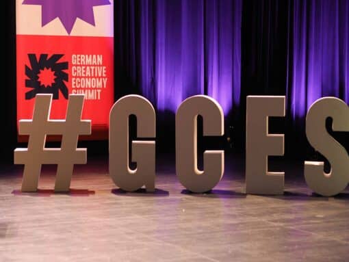 Das Bild zeigt große, dreidimensionale Buchstaben, die zusammen "#GCES" ergeben, auf einer Bühne mit einem violetten Vorhang im Hintergrund. Links im Bild ist ein Teil eines Plakats sichtbar mit der Aufschrift "GERMAN CREATIVE ECONOMY SUMMIT" neben einer stilisierten Blume in Rot und Lila.