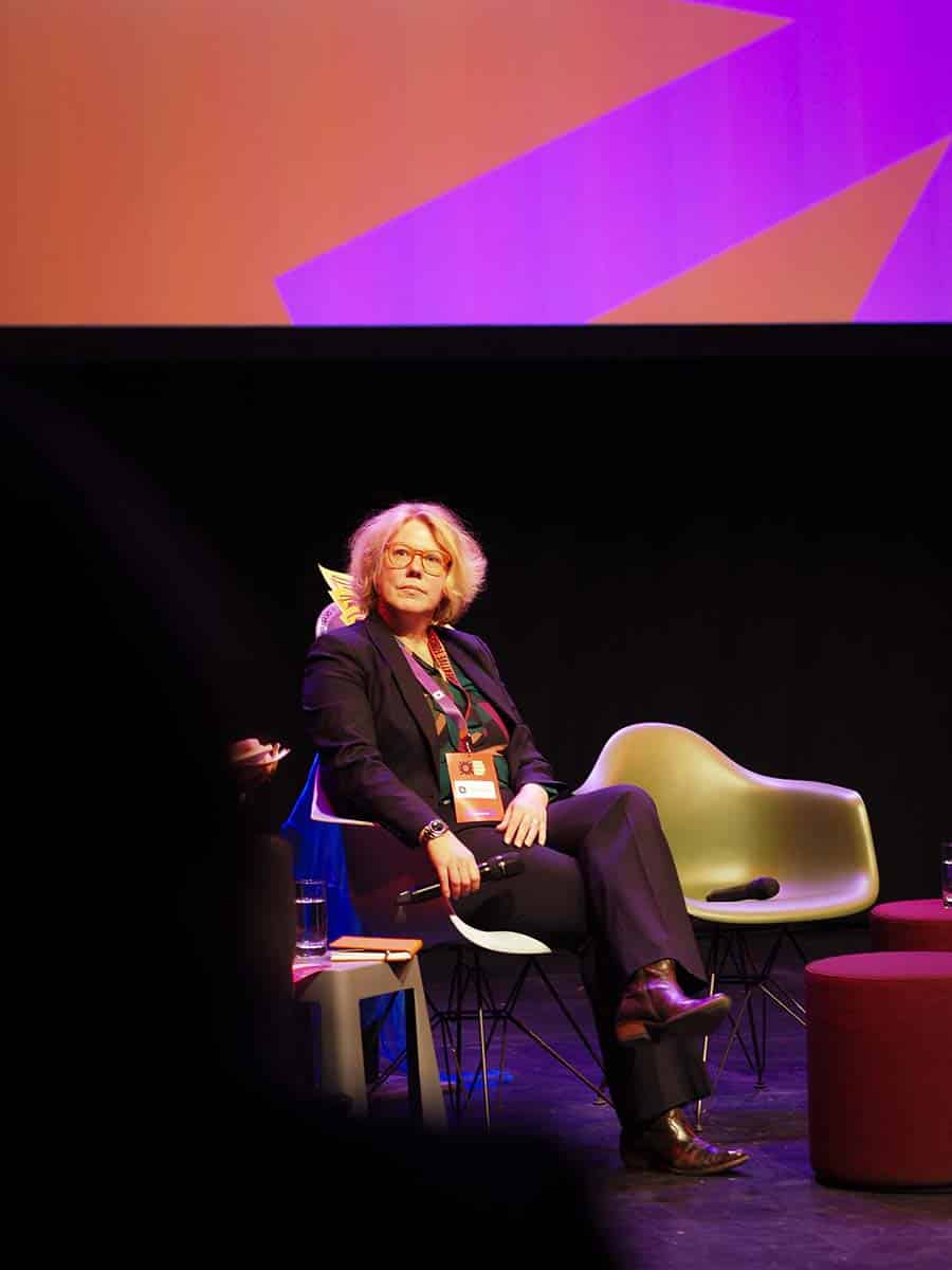 Eine Person mit blonden Haaren sitzt auf einem Stuhl auf einer Bühne, gestikuliert mit der Hand und spricht ins Mikrofon. Sie trägt einen dunklen Blazer und eine rote Akkreditierungskarte.