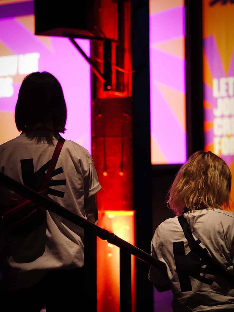 Zwei Personen von hinten, die auf einer Treppe stehen und auf eine Bühne mit leuchtenden Grafiken blicken. Die nähere Person trägt ein weißes T-Shirt mit einem schwarzen "KE" Symbol auf dem Rücken.