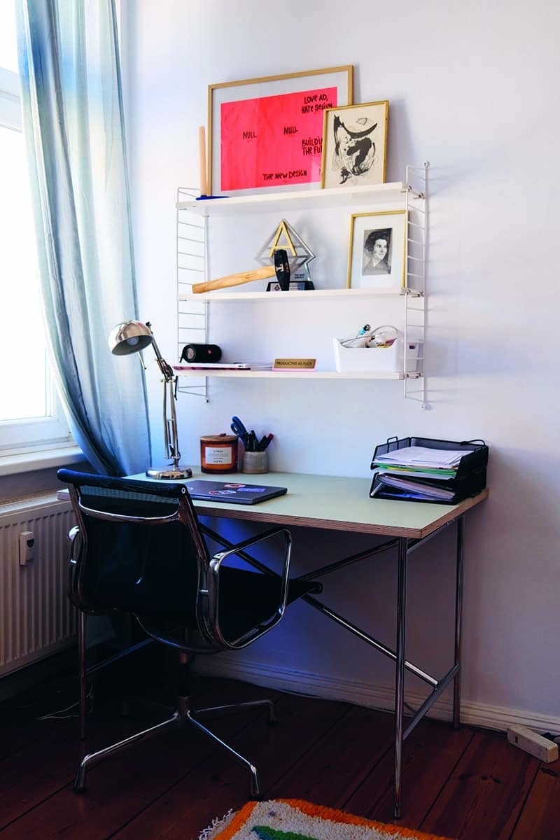 Arbeitsplatz mit Regalen und Dekoration: Ein moderner Arbeitsplatz mit einem weißen Schreibtisch, einem schwarzen Bürostuhl und weißen Regalen an der Wand. Auf den Regalen befinden sich Bücher, Kunstwerke und persönliche Gegenstände.