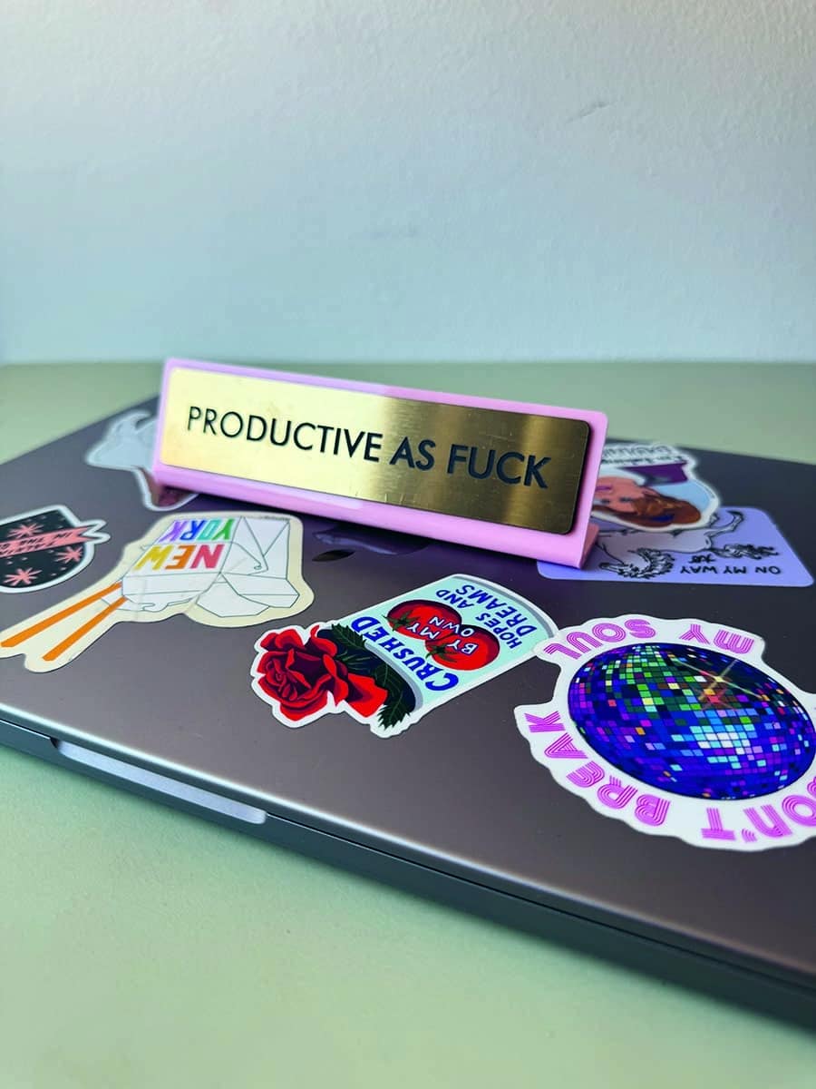 Laptop mit Motivationsschild und Stickern: Ein Laptop mit verschiedenen bunten Stickern beklebt. Auf dem Laptop steht ein Schild mit der Aufschrift "Productive as Fuck."