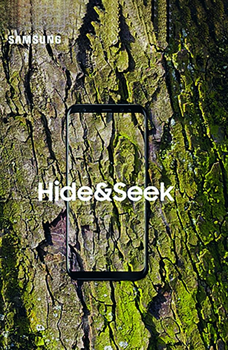 Samsung-Werbung mit "Hide & Seek" Text: Ein Samsung-Smartphone mit durchsichtigem Bildschirm, der sich nahtlos in die Textur eines Baumstamms einfügt. Der Text "Hide & Seek" ist auf dem Bildschirm zu sehen.