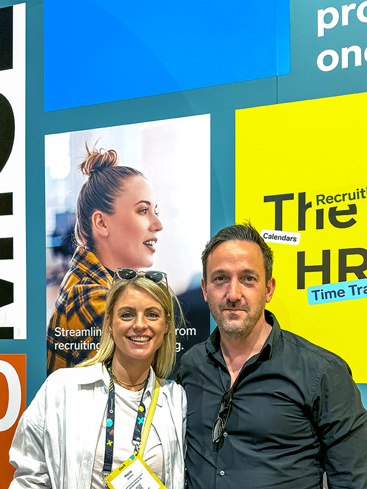 Foto eines lächelnden Mannes und einer Frau vor einem Messestand mit großen Werbetafeln und Firmenlogos