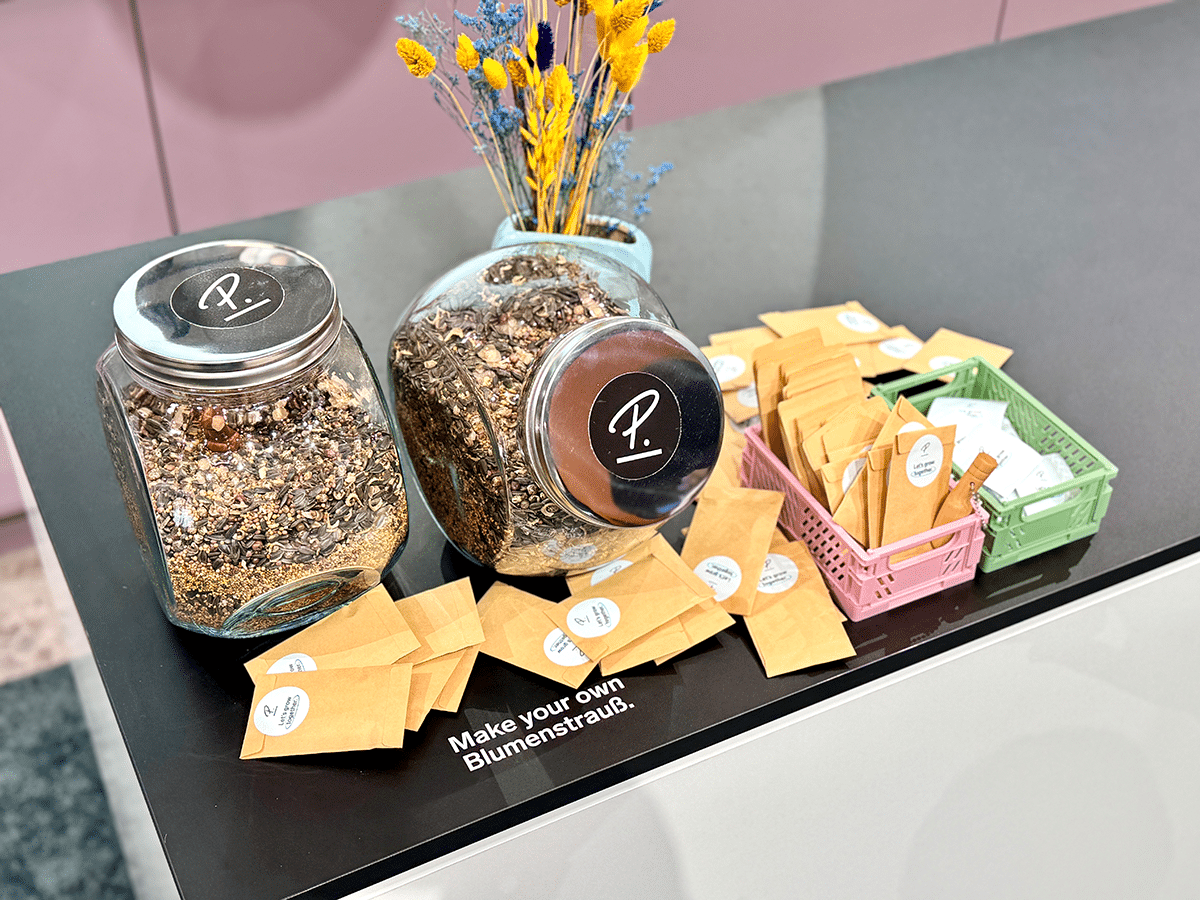 Nahaufnahme von Glasgefäßen mit Trockenblumen und Bastelmaterialien auf einem dunkelgrünen Tisch bei einer Messe.