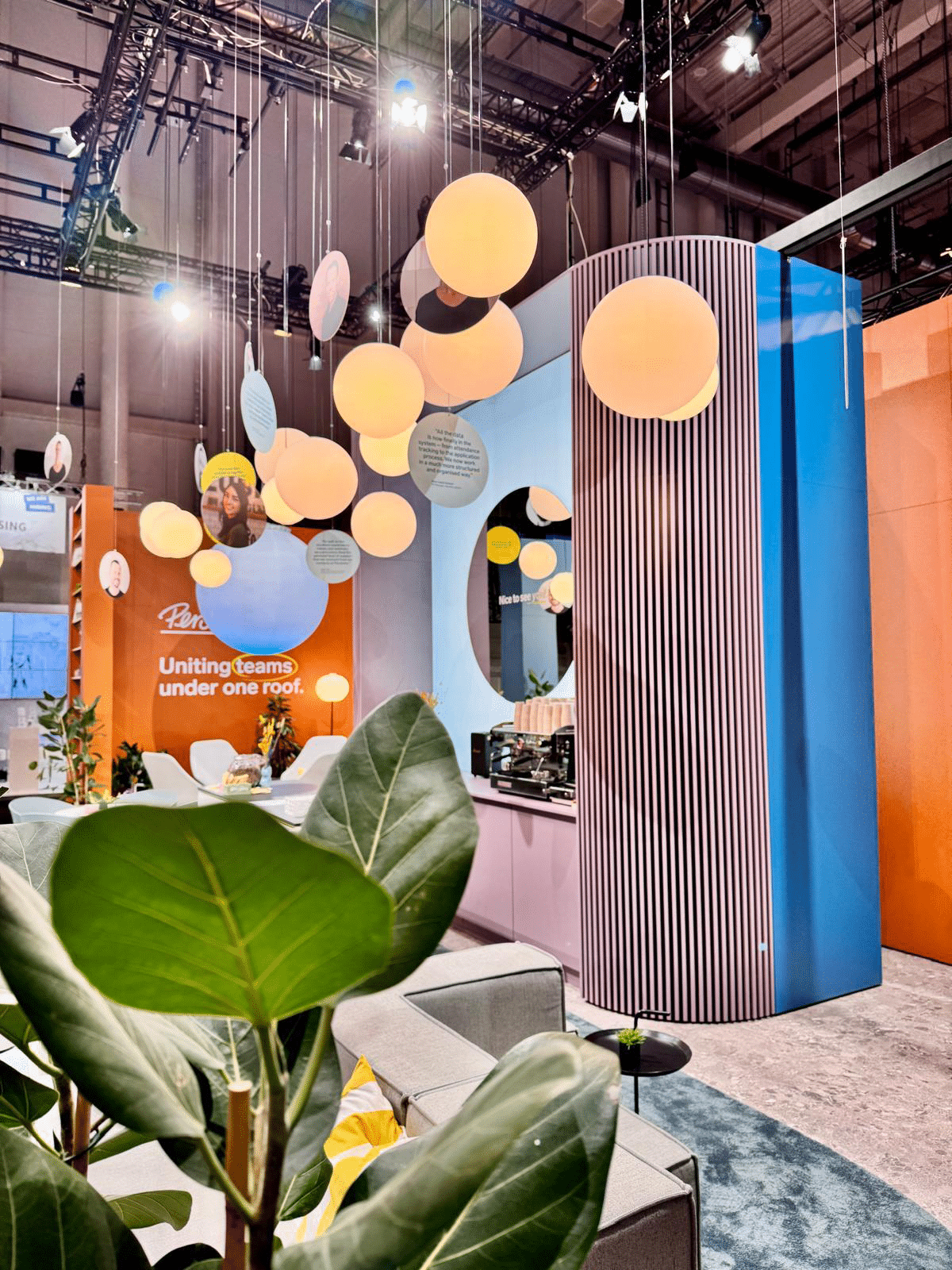 Detailansicht eines Messestands mit hängenden kreisförmigen Lampen, einer großen Pflanze im Vordergrund und einem hoch gestreiften Säulenelement