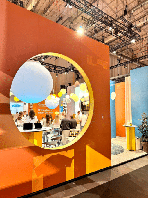 Durchblick durch einen großen, kreisförmigen, orangefarbenen Ausschnitt, der den Blick auf eine belebte Konferenzlounge freigibt.