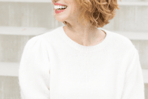 Porträt einer lächelnden Frau mit lockigem Haar, die ein weißes Oberteil trägt und auf Außentreppen sitzt