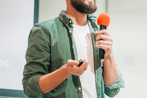 Ein kahlköpfiger Mann in einem grünen Hemd hält einen Vortrag. Er steht mit einem Mikrofon in der Hand und gestikuliert, während er neben einem Rednerpult steht