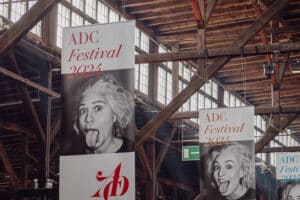Mehrere große Banner hängen von der Decke einer Veranstaltungshalle, die für das "ADC Festival 2024" werben. Auf den Bannern ist ein schwarz-weißes Bild einer Frau zu sehen, die ihre Zunge herausstreckt.