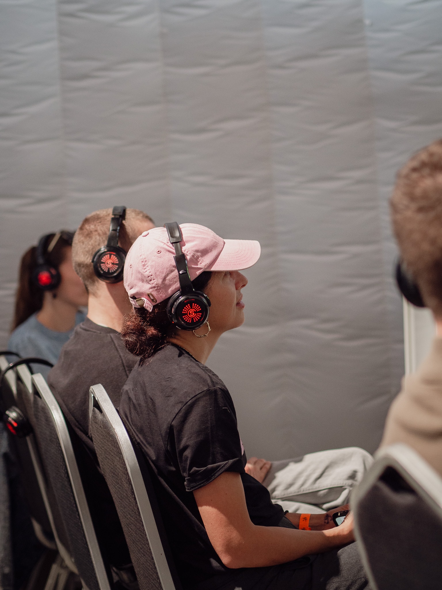 Mehrere Personen sitzen nebeneinander und tragen Kopfhörer, während sie einer Präsentation oder einem Vortrag zuhören. Im Vordergrund ist eine Frau mit einer rosa Kappe und einem schwarzen T-Shirt zu sehen.