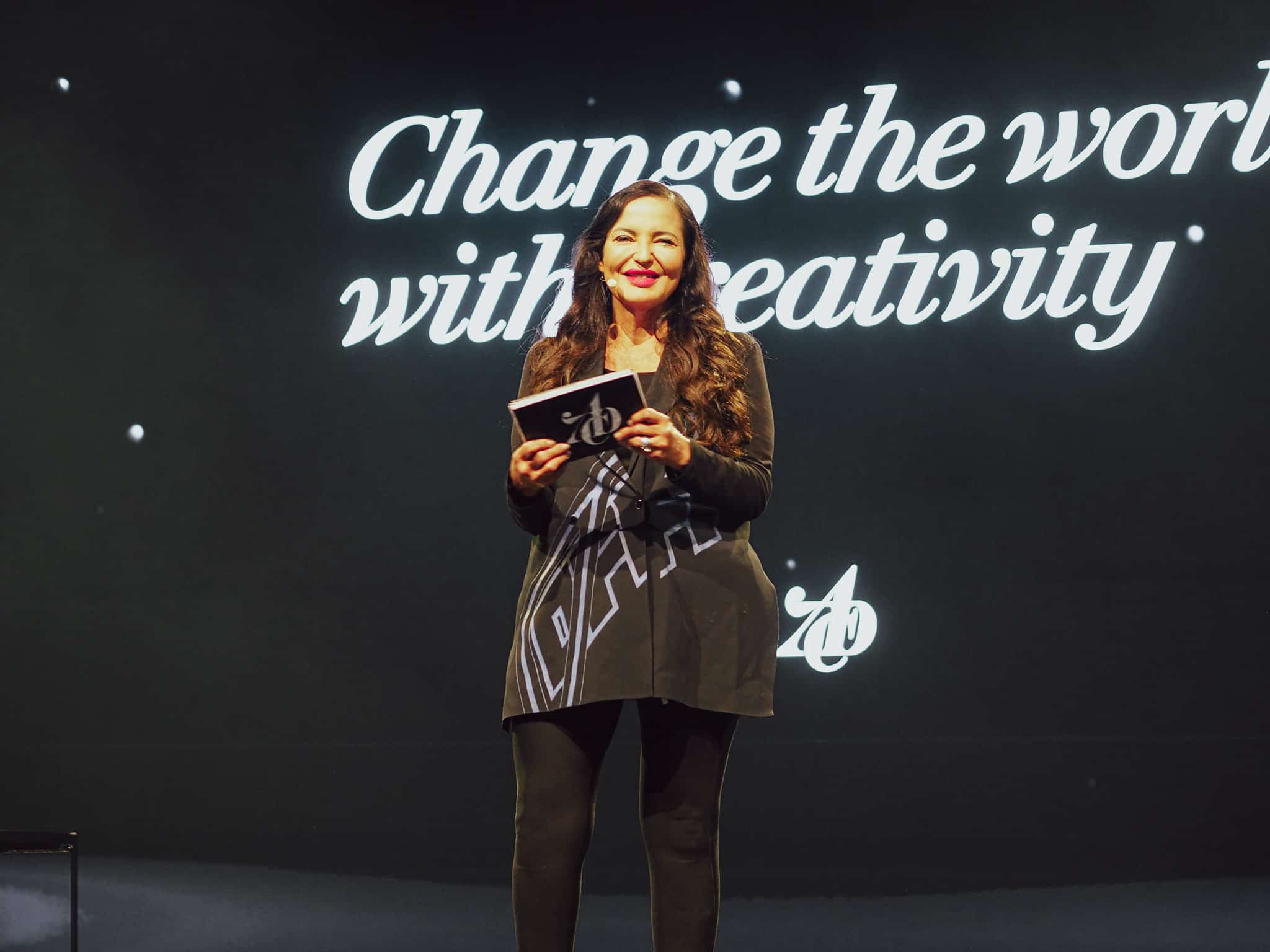 Eine Frau in schwarzer Kleidung steht auf einer Bühne und hält ein Mikrofon. Im Hintergrund befindet sich ein großer Bildschirm mit der Aufschrift "Change the world with creativity".