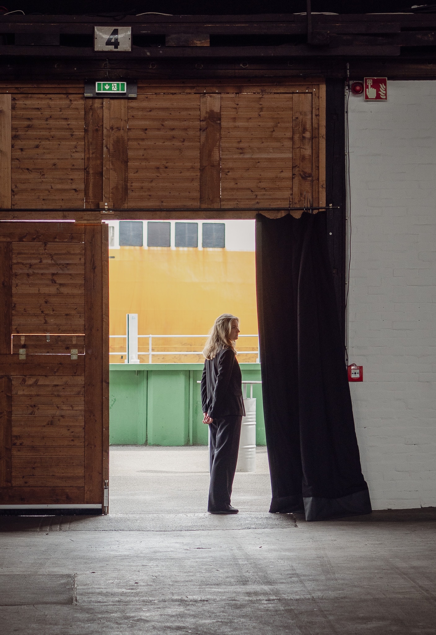 Eine Frau in schwarzer Kleidung steht in einer offenen Tür und blickt nach draußen, wo ein großes gelbes Schiff und ein grüner Container zu sehen sind.